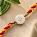 Close-up of a Gomti Chakra rakhi with red and yellow braided thread, featuring a white shell centerpiece and gold accents on a soft neutral background