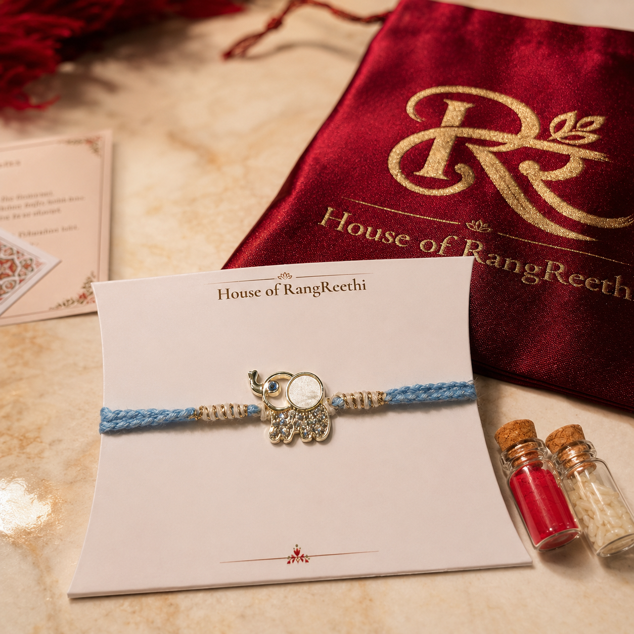 Blue braided baby rakhi with gold elephant charm displayed on a House of RangReethi card with red potli, roli chawal bottles, and Raksha Bandhan greeting cards.
