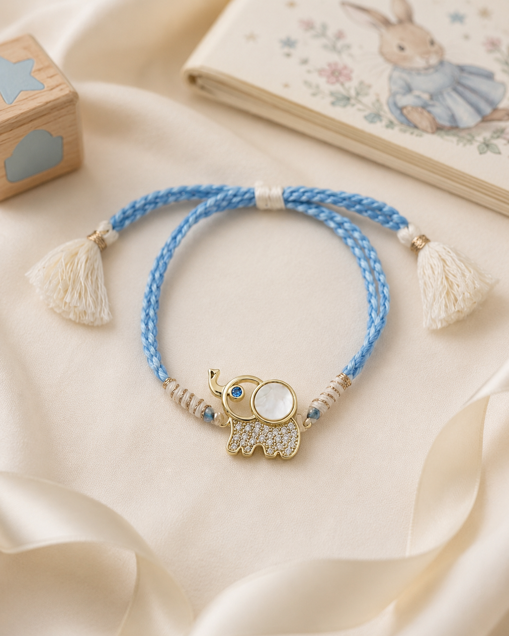 Blue braided baby rakhi with gold elephant charm, white tassel ends, pearl accents, and blue stones displayed on cream fabric with baby book and toy block in the background.