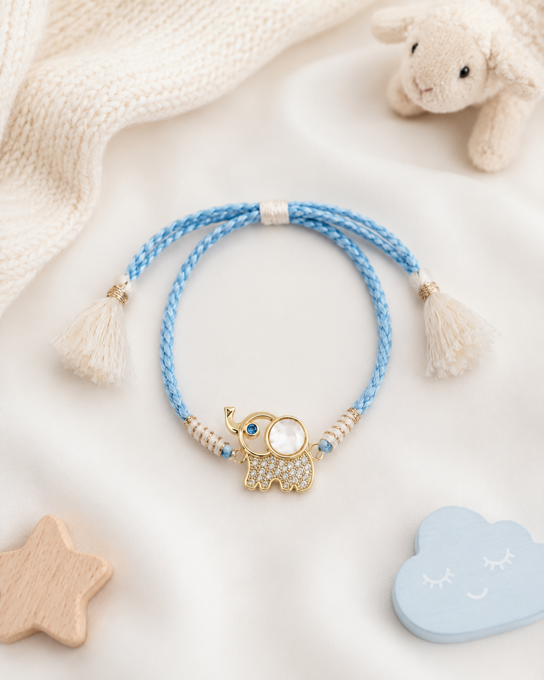 Blue braided baby rakhi with gold elephant charm, white tassel ends, and blue stone accents displayed on a soft white background with baby toys and plush decor.
