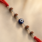 Close-up of an evil eye rudraksha rakhi with red thread, silver accents, and brown rudraksha beads on a warm neutral background.