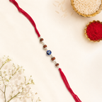 Evil eye rudraksha rakhi with red thread displayed on a cream background with rice, kumkum, and white flowers.