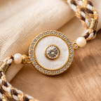 Close-up of a gold and ivory rakhi with mother of  pearl centerpiece, braided thread on beige fabric and wood background.