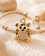 Close-up of a gold teddy bear baby rakhi with beige thread, tassel ends, and crystal detailing on a soft cream fabric background.