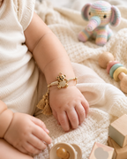 Gold teddy bear baby rakhi with beige thread and tassel ends tied on a baby’s wrist, shown on soft cream fabric with baby toys in the background