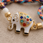 Close-up of a multicolor braided rakhi with white elephant charm, gold detailing, colorful stones, and pearl accents on a beige fabric background.