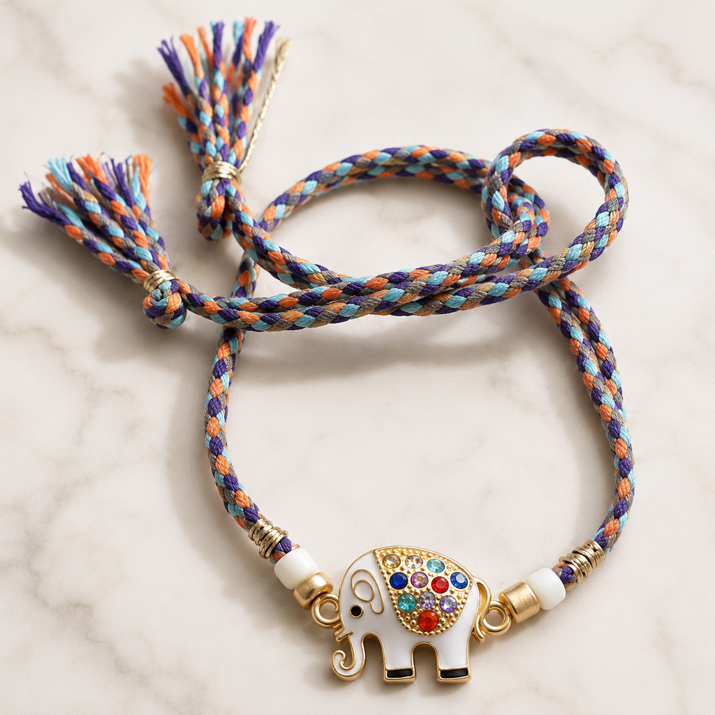 Multicolor braided rakhi with white elephant charm, colorful stone detailing, gold accents, pearl beads, and tassel ends displayed on a white marble background.