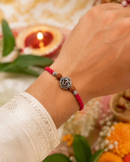 Om rudraksha rakhi with red thread tied on a wrist, styled with festive diya, flowers, and traditional Raksha Bandhan decor.