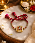Red braided rakhi with gold square Jadau Kundan centerpiece, pearl accents, and tassel ends displayed on a marble surface with diya, roli, and chawal in the background.