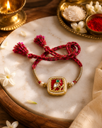 Red braided rakhi with gold square Jadau Kundan centerpiece, pearl accents, and tassel ends displayed on a marble surface with diya, roli, and chawal in the background.
