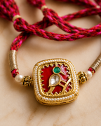 Close-up of a red braided rakhi with gold square Jadau Kundab centerpiece, pearl accents on a marble background.