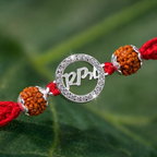 Close-up of a silver Ram rudraksha rakhi with red thread, brown rudraksha beads, silver accents, and crystal detailing on a green leaf background.