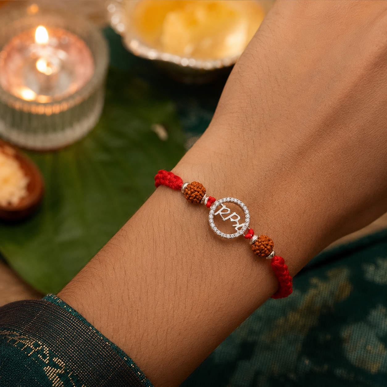 Silver Bhai rudraksha rakhi with red thread tied on a wrist, styled with a diya, green leaf, and festive Raksha Bandhan decor.