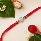 Silver Bhai rudraksha rakhi with red thread displayed on a cream background with kumkum, rice, wooden spoons, and green leaves