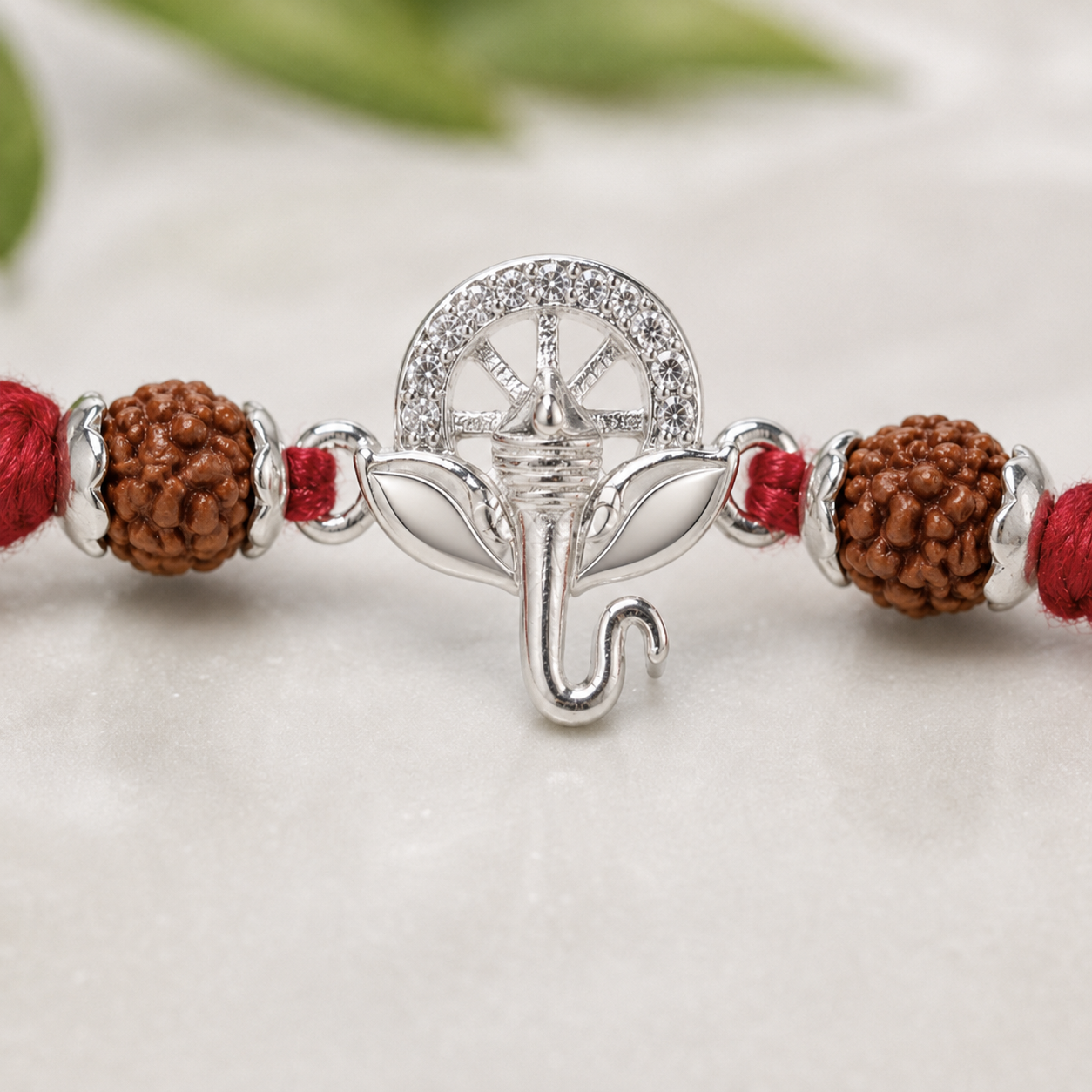 Close-up of a silver Ganesh rudraksha rakhi with red thread, featuring brown rudraksha beads and silver accents on a soft neutral background.