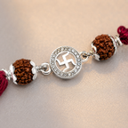 Close-up of a silver Swastik rudraksha rakhi with red thread, brown rudraksha beads, silver accents, and crystal detailing on a soft neutral background.