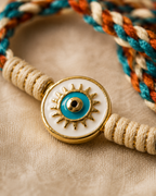 Close-up of a turquoise evil eye rakhi with gold sunburst detailing, cream thread wrapping, and teal orange braided thread on a beige fabric background.