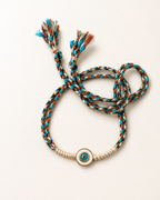 Turquoise, brown, and cream braided evil eye rakhi with tassel ends and gold-accented centerpiece displayed on a clean white background.