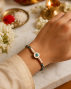Turquoise and gold evil eye rakhi tied on a wrist with cream thread wrapping, shown with diya, roli, chawal, and jasmine flowers in the festive background.