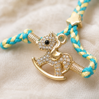 Close-up of a turquoise and yellow braided baby rakhi with gold rocking horse charm, crystal detailing, and star accent on a soft cream fabric background.