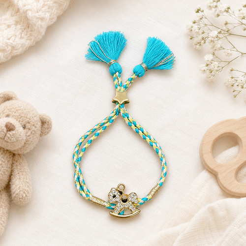Turquoise and yellow braided baby rakhi with gold rocking horse charm, star accent, and tassel ends displayed on soft cream fabric with baby toys and flowers.